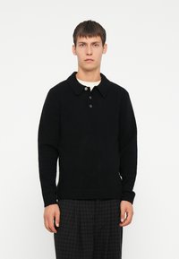 Schwarzer strukturierter Strickpullover mit Kragen, ausgestattet mit einer Knopfleiste und gerippten Bündchen. Getragen mit karierter Hose.