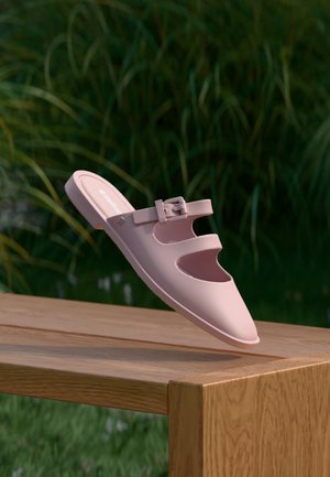 Light pink slip-on sandal with two straps and a buckle, floating above a wooden bench with green foliage in the background.