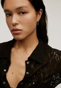 Camisa de encaje negra con collar, adornada con botones plateados y una textura transparente, combinada con pequeños aros en las orejas. Retrato en primer plano.