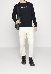 Felpa nera con logo "REPLAY", pantaloni bianchi e stivali neri con lacci. Modello tiene una borsa a tracolla verde scuro. Sfondo semplice.