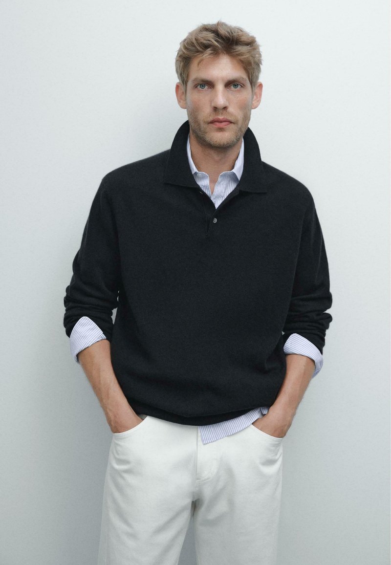 Schwarzer Pullover mit Kragen, hochgekrempelte Ärmel, die ein blau-gestreiftes Hemd darunter zeigen, getragen mit weißen Hosen. Einfarbiger Hintergrund.