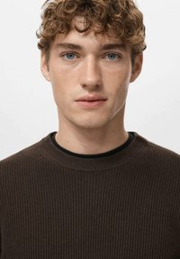 Brauner, gerippter Pullover mit rundem Ausschnitt, kombiniert mit einer schwarzen Innenschicht. Das Modell hat lockiges Haar und blaue Augen und schaut direkt in die Kamera.