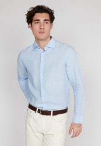 Chemise en lin bleu clair à manches longues avec un col pointu et des boutons blancs, associée à un pantalon blanc et une ceinture en cuir brun.