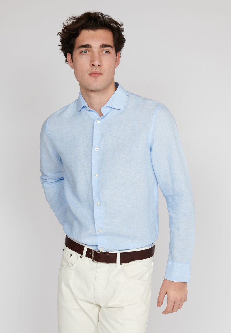 Chemise en lin bleu clair à manches longues avec un col pointu et des boutons blancs, associée à un pantalon blanc et une ceinture en cuir brun.