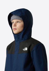 Wodoodporna kurtka w granatowym kolorze z czarną panelem na piersi, regulowanym kapturem i białym logo. Charakteryzuje się gładką teksturą i casualowym wzornictwem.