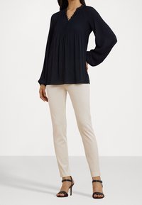 Kvinna som bär en lös svart långärmad blus, slim fit beiga byxor och svarta högklackade sandaler, poserar med ena handen på höften.