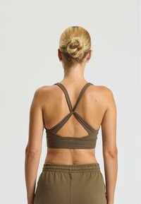 Reggiseno sportivo marrone con design incrociato a fascia sulla schiena, realizzato in materiale morbido ed elastico. Si abbina a pantaloni a vita alta color verde oliva.