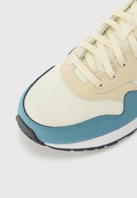 Sneaker mit einem hellgelben Mesh-Obermaterial, blauen synthetischen Überzügen, cremefarbenen Schnürsenkeln und einer strukturierten schwarzen Gummisohle. Abgerundete Zehenform.