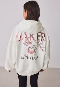 Balta didelio dydžio Džemperis su gaubtu, puoštas metalinėmis rožinėmis raidėmis, drugelio grafika ir dekoratyviniais elementais, su trikotažiniu apvadu apačioje ir rankogaliuose.