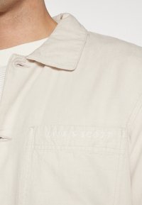 Lichtbeige jack van katoen met een kraagontwerp. Heeft een voorzak en een geborduurd "Lyle & Scott" logo in het wit.