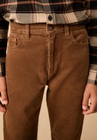Pantalon en velours côtelé marron avec une texture côtelée, des poches avant et une fermeture à bouton unique, associé à une chemise en flanelle à carreaux.