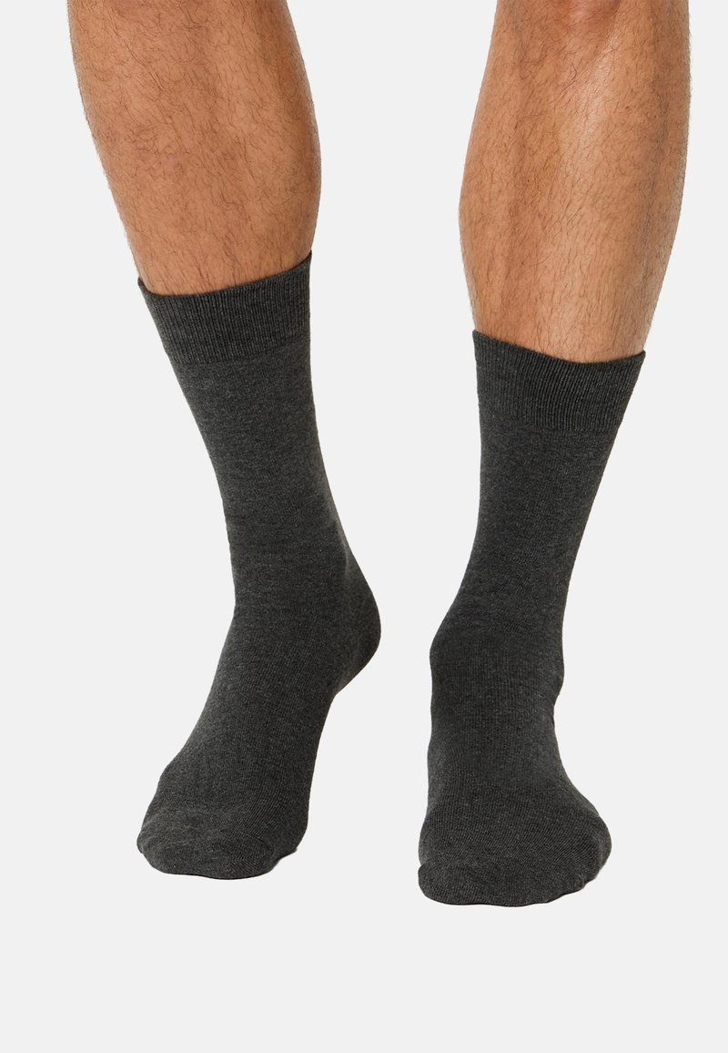 Nogi w ciemnoszarych skarpetach typu crew, stojące na jasnym tle z lekko rozstawionymi stopami i palcami skierowanymi do przodu.