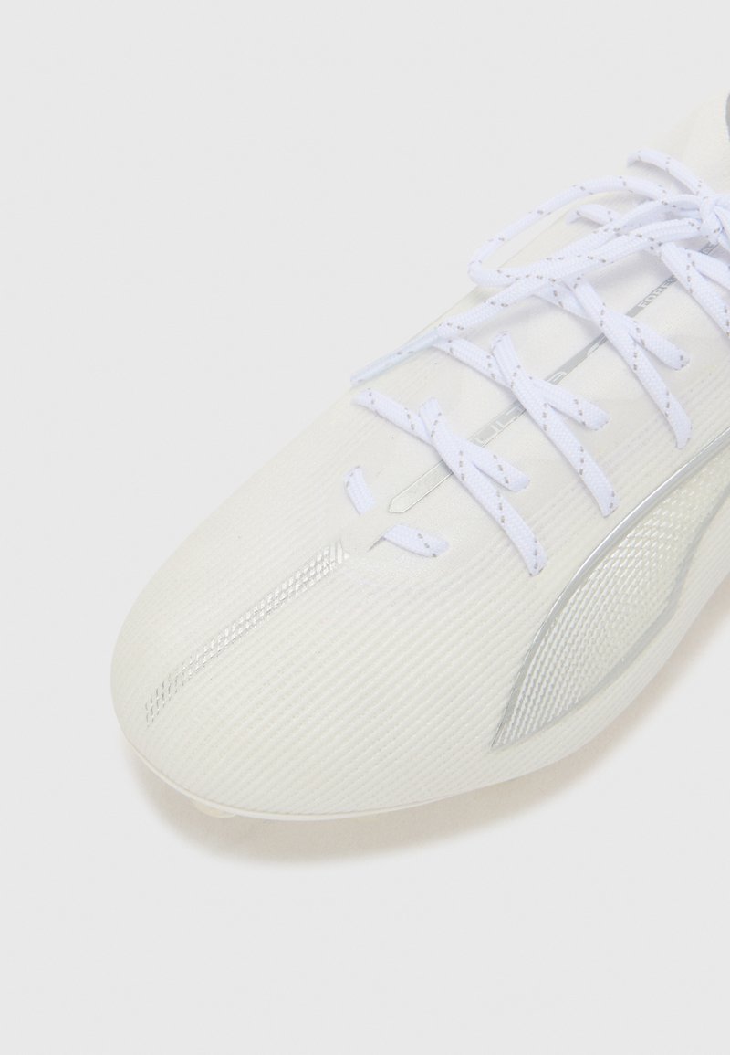 Zapatilla deportiva blanca con un upper texturizado, punta curvada y acentos reflectantes. Presenta cordones blancos y un diseño aerodinámico.
