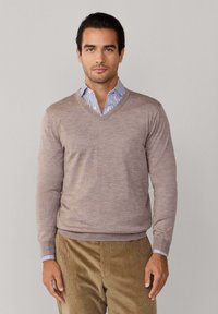 Svetlono hnedý sveter s výstrihom do v a mäkkou textúrou, nosený cez modro-bielu pruhovanú košeľu s golierom, s priliehavými rukávmi a rebrovaným lemom.