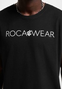 Černé bavlněné tílko bez rukávů s potiskem "ROCA WEAR" v bílé barvě. Výstřih ve tvaru kruhu s hladkou texturou a bez viditelných vzorů.