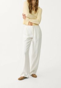 Lange witte broeken met een hoge taille, gecombineerd met een geribbelde, knoopsluiting, lichtgele top. Bruine mules versierd met zilveren studs maken de look compleet.