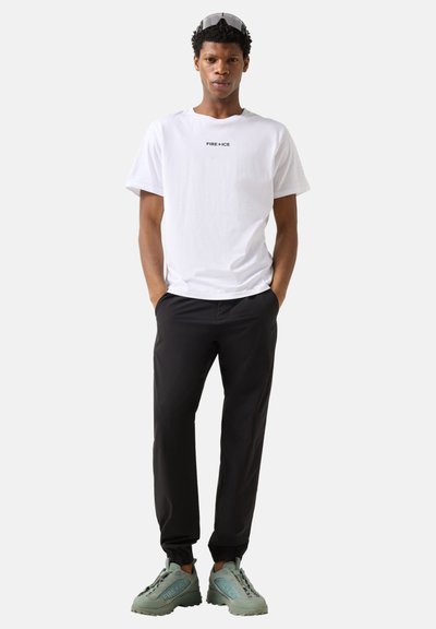 Homme portant un t-shirt blanc "FIRE + ICE", un pantalon noir, des baskets épaisses gris-vert et des lunettes de soleil sur la tête, debout les mains dans les poches.