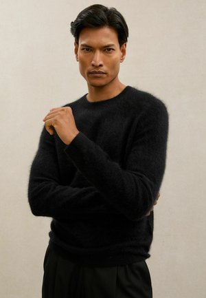 Schwarzer flauschiger Pullover mit rundem Ausschnitt, gerippten Bündchen und weicher Textur. Das Model posiert mit gekreuzten Armen vor einem neutralen Hintergrund.