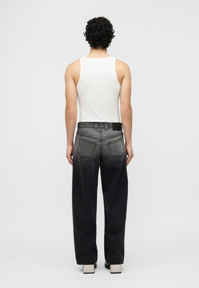 Άτομο με σγουρά σκούρα μαλλιά στέκεται πλάτη προς το φακό, φορώντας λευκό αμάνικο τοπ, ξεθωριασμένο μαύρο φαρδύ τζιν και λευκά παπούτσια πλατφόρμα.