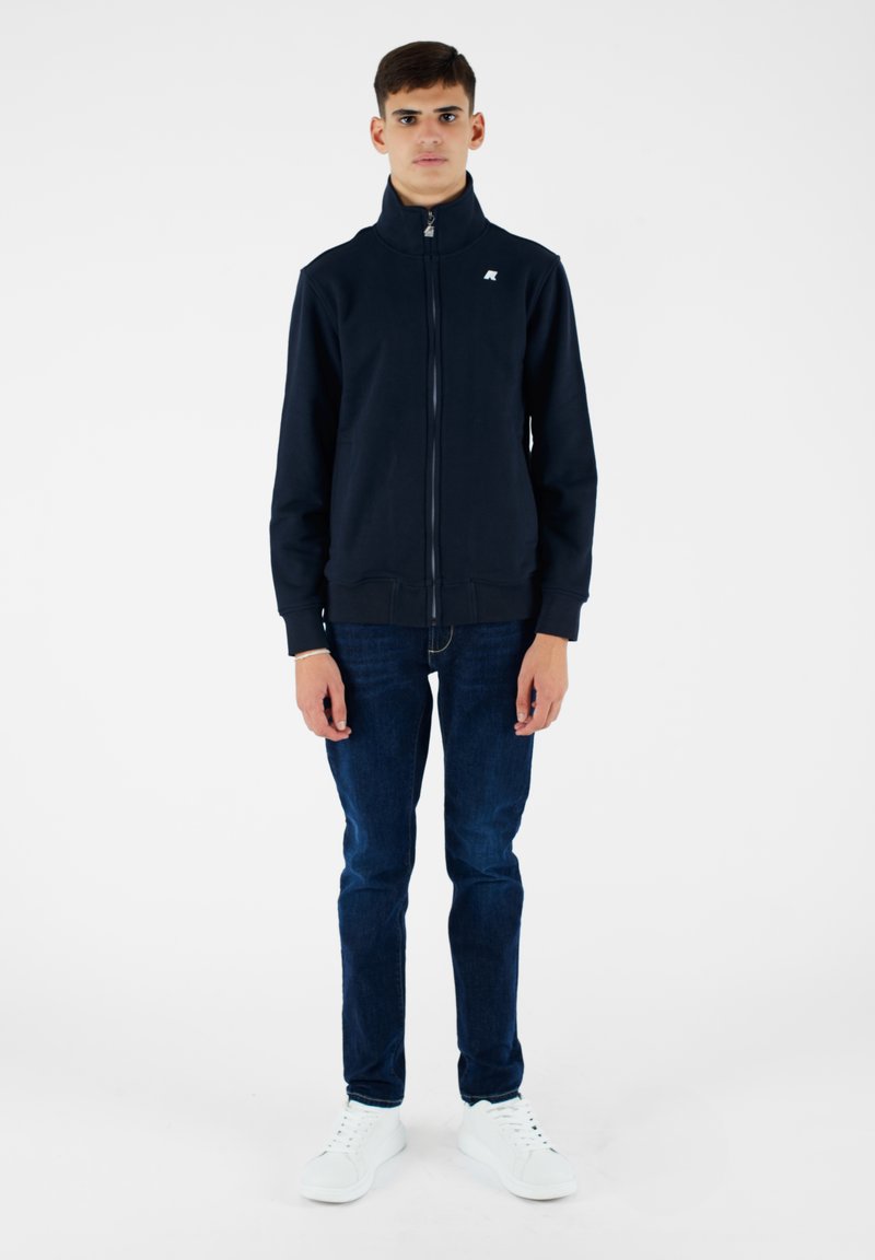 Giacca di jeans navy con zip e colletto alto, realizzata in tessuto morbido. Abbinata a jeans scuri e sneaker bianche, crea un look casual.