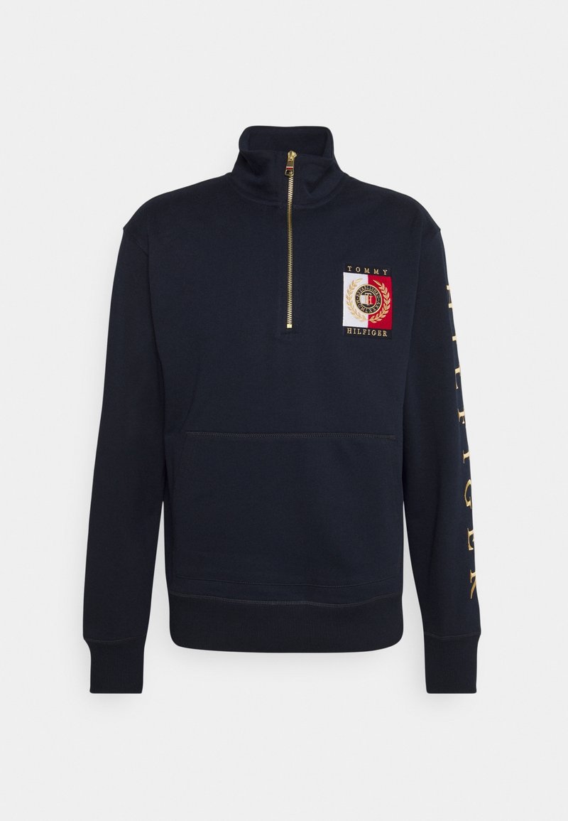 Sweat-shirt bleu marine à fermeture éclair partielle avec un patch logo Tommy Hilfiger sur la poitrine et un lettrage doré sur la manche droite.