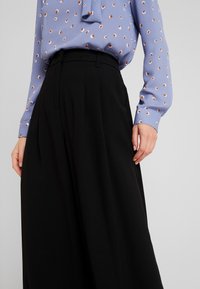 Kvinna klädd i svart byxor med hög midja och vida ben, samt en ljusblå blus med orange och vit prickmönster, stående med avslappnade händer.