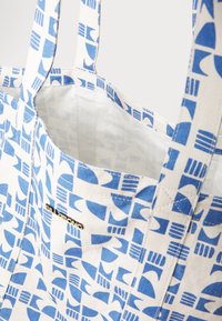 Canvas tote bag featuring a geometric blue and white pattern, two sturdy handles, and a gold logo accent near the opening.