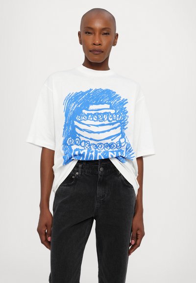 Balti per didelis T-shirt su mėlynu iliustruotu torto grafiniu atvaizdu priekyje, derinamas su juodais džinsais. Lygus audinys, atsitiktinis dizainas.