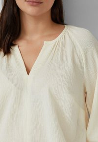 Helle beige Bluse mit plissierter Textur, V-Ausschnitt und gerafften Schultern. Der Stoff ist weich und leicht zerknittert.