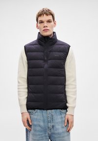 Melna pūkaina veste ar augstu apkakli, kvadrātveida rakstu, priekšējo rāvējslēdzēju un logotipu krūtīs, valkāta virs krēmkrāsas ribota džempera un zilām džinsa biksēm.