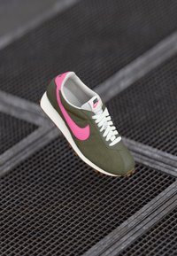 Olīvu zaļas sporta kurpes ar rozā Nike zīmi, baltām auklām un teksturētu virsējo daļu. Iekļauj baltu vidussole un gumijas zoli, kas nodrošina saķeri.