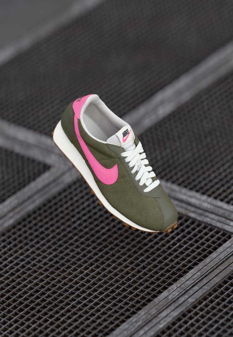 Olīvu zaļas sporta kurpes ar rozā Nike zīmi, baltām auklām un teksturētu virsējo daļu. Iekļauj baltu vidussole un gumijas zoli, kas nodrošina saķeri.