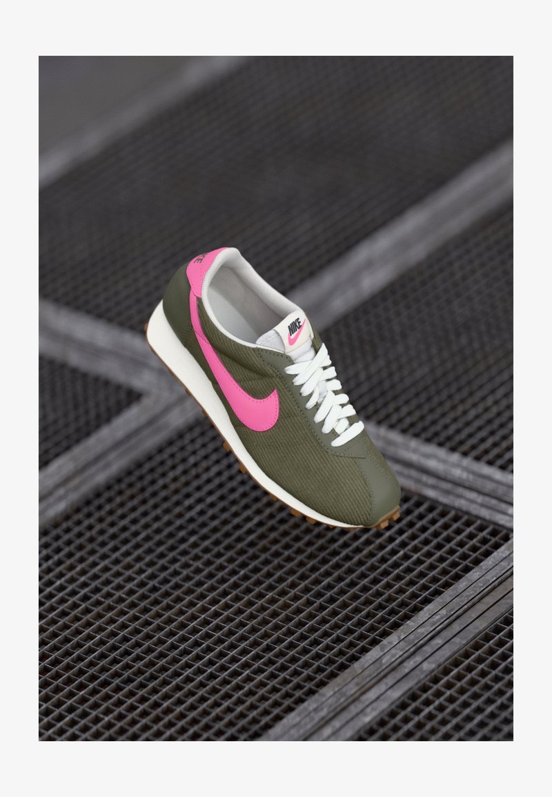 Olivnozeleni tenisice s roza Nike logotipom, belimi vezalkami in strukturirano zgornjo plastjo. Imajo belo srednjo podnožje in gumijast podplat za oprijem.