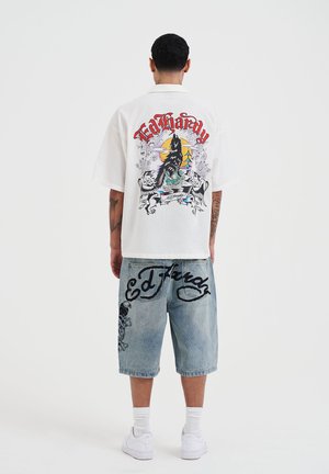 Camiseta blanca de gran tamaño con un diseño gráfico colorido en la parte posterior, combinada con pantalones cortos de mezclilla azul claro que presentan un logotipo negro y acentos artísticos.