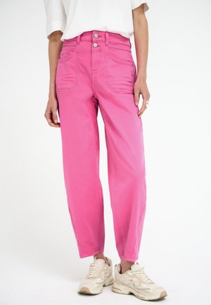 Personne portant un pantalon rose taille haute avec deux poches avant et des baskets blanches, debout sur un fond blanc uni.