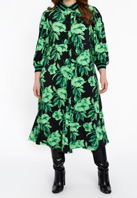 Musta mekko, jossa on eläväinen vihreä kukkakuosi, nappikiinnitys ja kaulus, jossa on vihreä- ja mustaraitaiset yksityiskohdat hihoissa.
