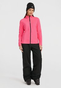 Fleece jaka spilgti rozā krāsā ar augstu apkakli un priekšējo rāvējslēdzēju, savienota ar melniem kravas biksēm un melniem sniega zābakiem.
