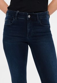 Jeans in denim blu scuro con un design aderente, vita alta, tasche anteriori e dettagli in bottone argentato. Tessuto liscio con minima scoloritura.