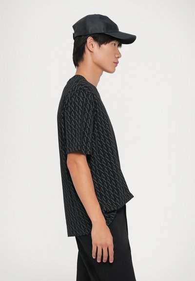 Un t-shirt noir à manches courtes présente un motif intégral de lettres stylisées. Le modèle porte une casquette noire avec une finition brillante.