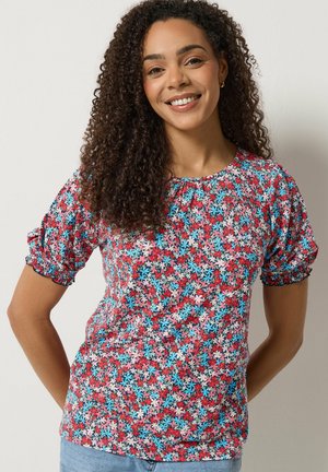 Femme aux cheveux bouclés portant un haut à manches courtes coloré avec des fleurs rouges, bleues et roses, souriant sur un fond clair.