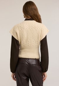 Kremowy sweter z dzianiny z teksturowanym wzorem, ściągaczem w talii i krótkimi rękawami, połączony z ciemnobrązową bluzką oraz brązowymi skórzanymi spodniami.