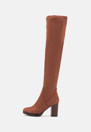 Bottes en daim marron montant jusqu'aux genoux avec un talon bloc. Elles présentent une découpe en forme de V sur les côtés et une texture lisse.