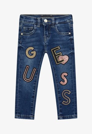 Jean en denim bleu foncé pour enfants avec des lettres ornées de sequins formant le mot « GUESS » et un cœur en sequins sur les jambes avant.