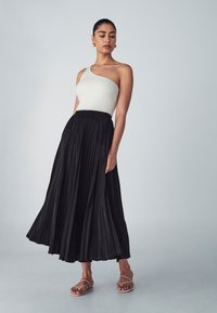 Black pleated maxi skirt with a high waistband, paired with a white ribbed one-shoulder top. Simple flat sandals complete the look.