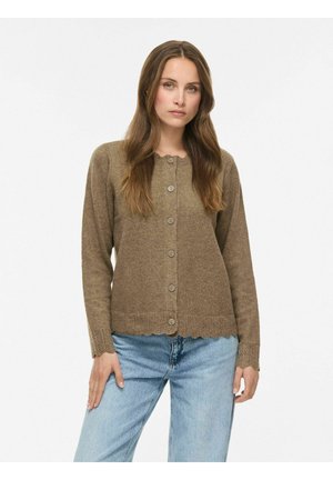 Femme aux longs cheveux bruns portant un cardigan marron boutonné et un jean bleu clair sur un fond blanc uni.