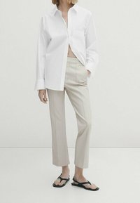 Chemise blanche à manches longues avec une poche avant, associée à un pantalon beige sur mesure et des tongs noires. Coupe décontractée et confortable. Texture lisse.