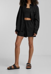 Personne portant un crop top noir, une chemise ample noire à boutons, un short noir à taille élastique et des sandales à plateforme noires, se tenant devant un fond uni.