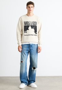 Young man wearing beige Nørgaard sweatshirt with black graphic and text, ripped blue jeans, and white sneakers, standing against a plain background.
