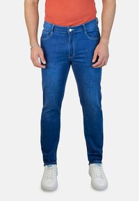Jeans de mezclilla azul con un corte slim, con cierre de botón en la parte delantera, cinco bolsillos y costuras en contraste. Combinados con zapatillas blancas.