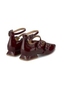 Chaussures en cuir verni bordeaux avec des talons crantés, ornées de sangles décoratives et d'accents en gemmes, alliant texture brillante et design structuré.